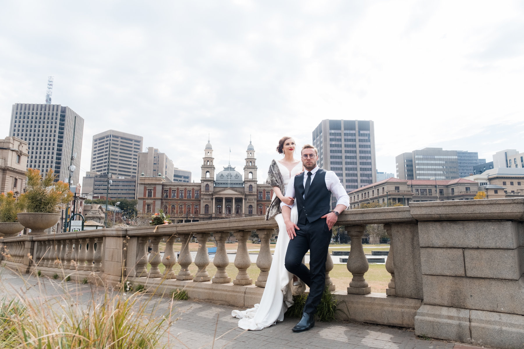 Wide angle view of beautiful, old buildings and railings with bride and groom together, looking on.